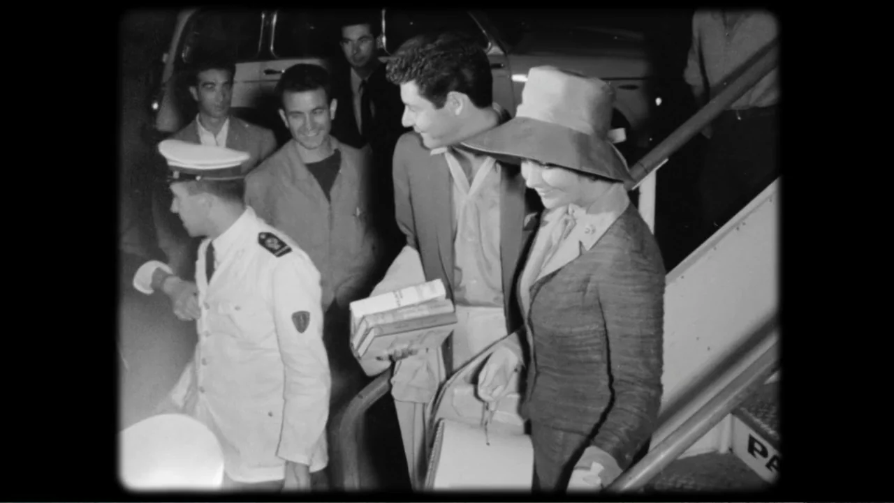 Elizabeth Taylor: The Lost Tapes (2024) movie still: medium — A group of people descend outdoor airport stairs at night near an aircraft fuselage. A…
