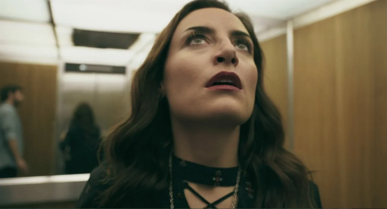 The Lift (2021) movie still: closeup — A small elevator with light wood paneled walls is illuminated by bright overhead lights. A woman with long…
