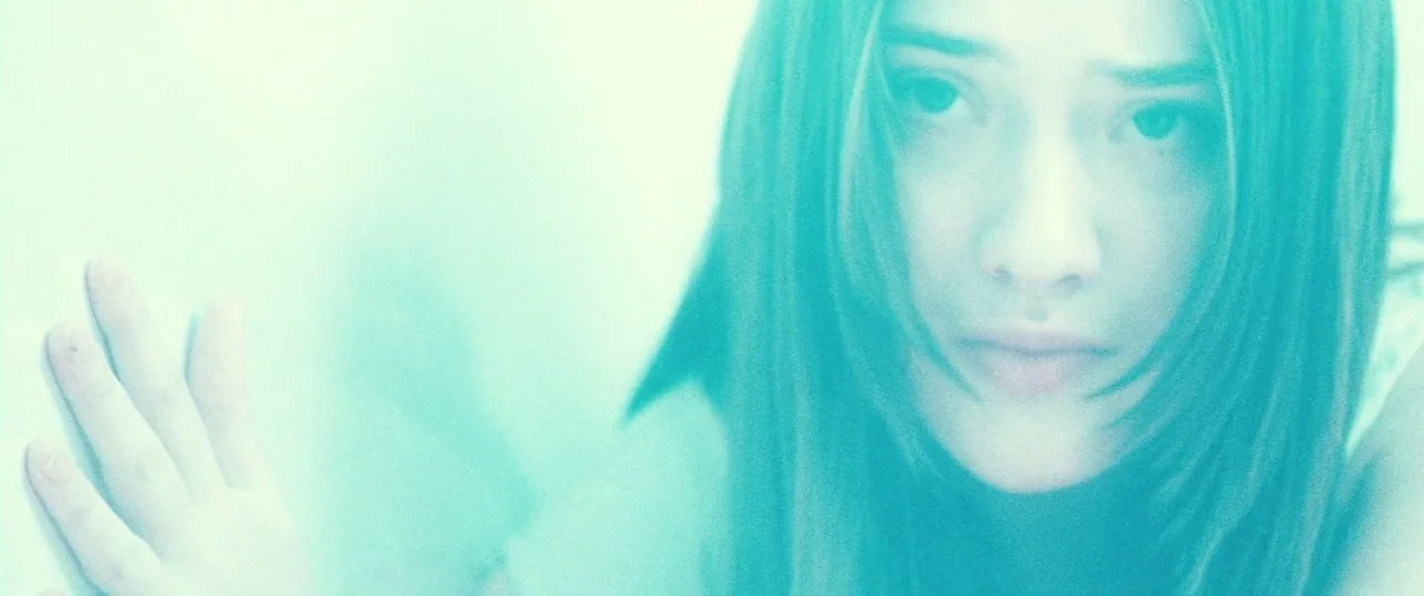 Beyond The Black Rainbow (2010) movie still: closeup — A close-up view of a woman against a bright, featureless surface. Her hand touches the wall…