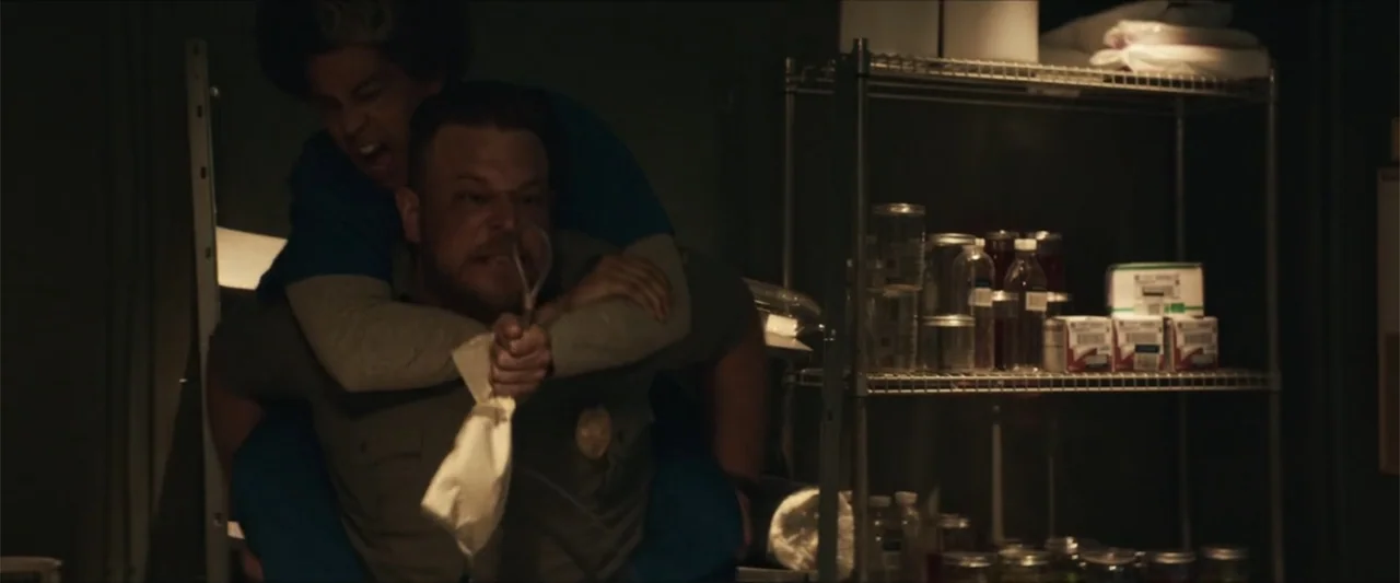 American Carnage (2022) movie still: medium — A dark storage room contains industrial wire shelving units filled with various jars and boxes. A…
