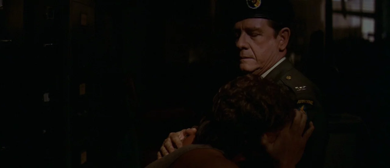 First Blood (1982) movie still: medium — A dimly lit interior space with rows of metal filing cabinets visible in darkness. An older man in uniform…