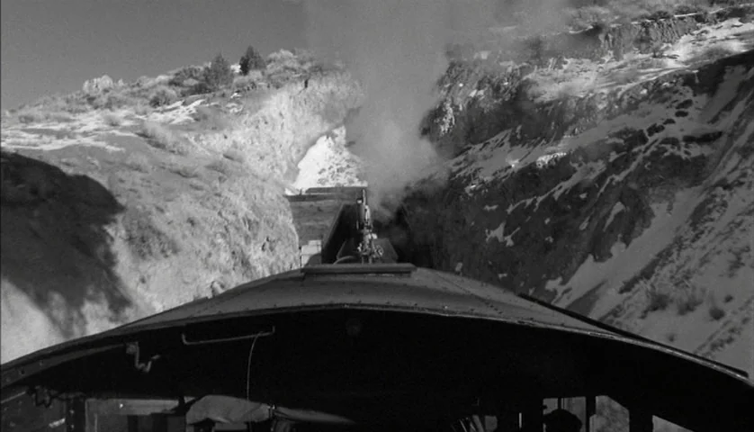 Dead Man (1995) movie still: wide — A steam train moves through a craggy, snow-dusted mountain canyon under bright daylight. Thick smoke rises from…