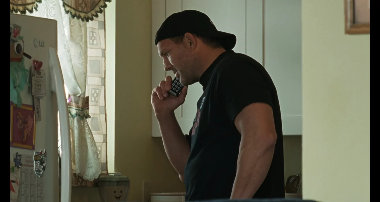 The Smashing Machine (2025) movie still: medium — A man stands in a residential kitchen with white cabinets and a window. The man holds a telephone…