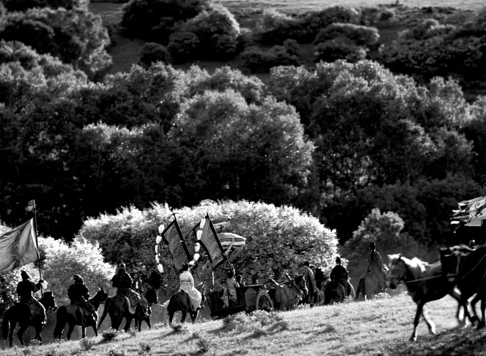The Assassin (2015) movie still: wide — A group of people on horseback follow a horse-drawn cart across a grassy hill. The background is filled with…