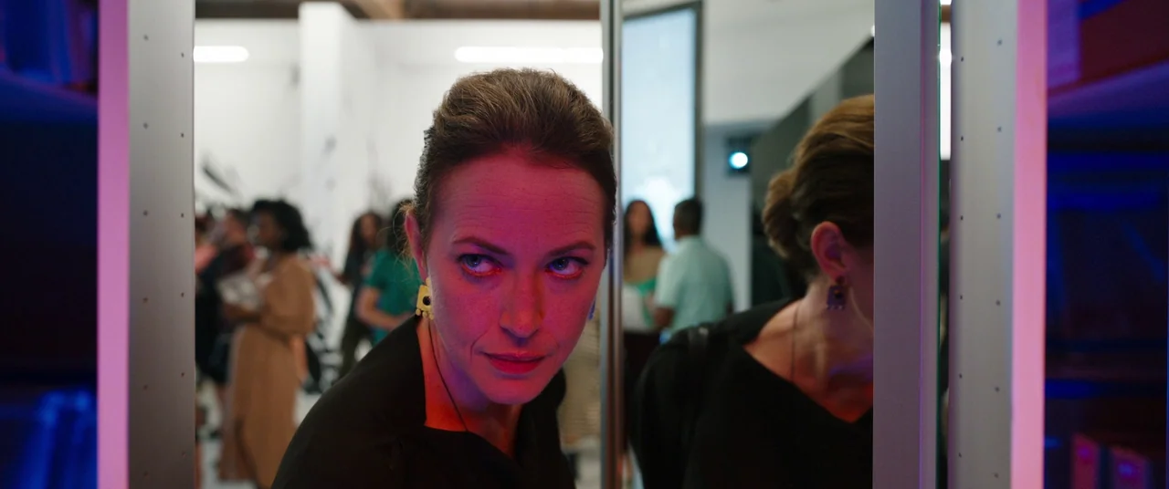 Candyman (2021) movie still: closeup — An interior space with multiple visitors blurred in the background of a brightly lit area. A woman looks…