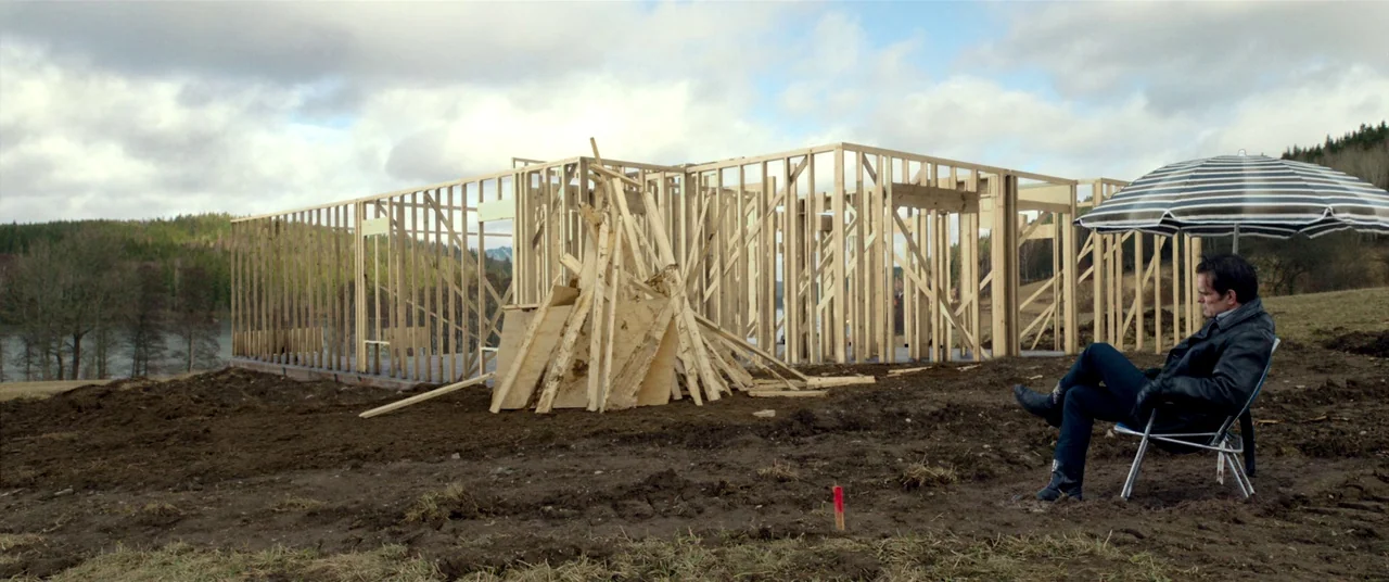 The House That Jack Built (2018) movie still: wide — A field contains an unfinished wooden house frame next to a lake. A man sits in a folding chair…