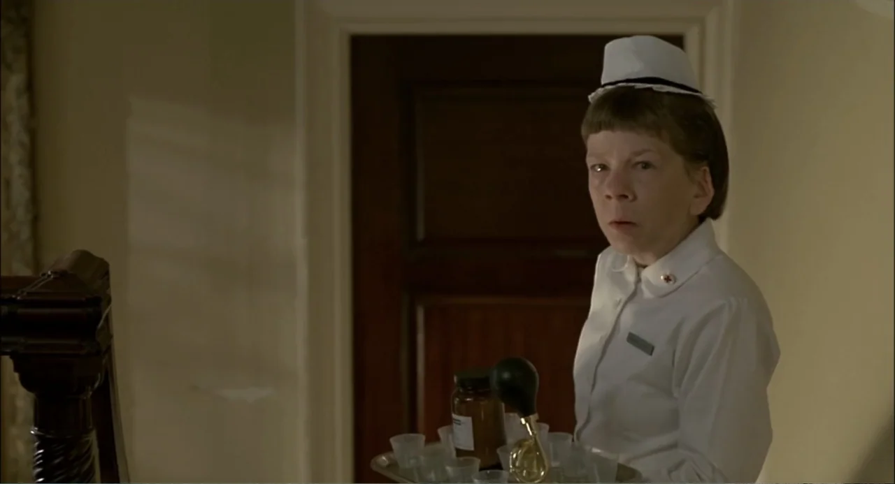 She-Devil (1989) movie still: medium — An interior hospital corridor with plain beige walls and a dark wooden door. A nurse in a white uniform holds…