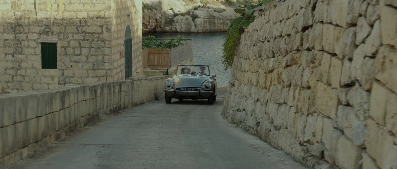 By The Sea (2015) movie still: wide — A narrow road lined with rough stone walls leads toward a bay of water. A vintage convertible car drives up…