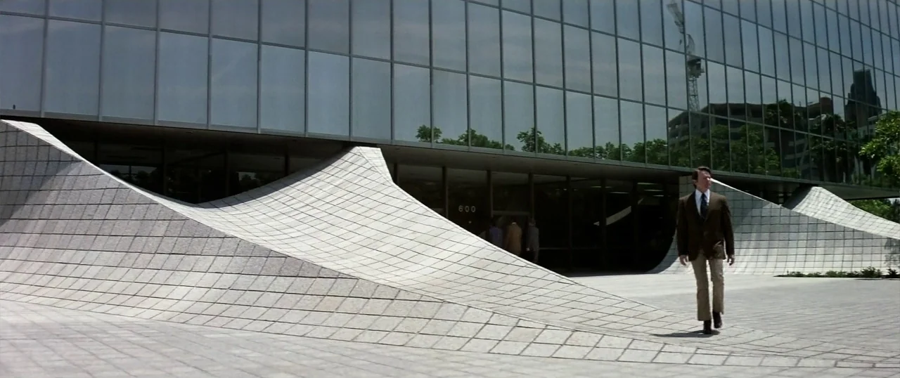 The Parallax View (1974) movie still: fullbody — A modern plaza features curved white stone tile structures and a glass-paneled building facade. A…