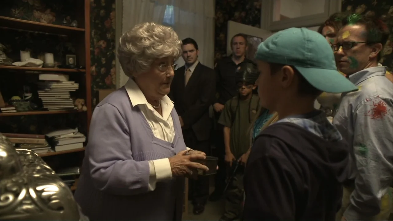 Boathouse Detectives (2010) movie still: medium — A cluttered residential living room with floral wallpaper and a wooden shelving unit. An elderly…