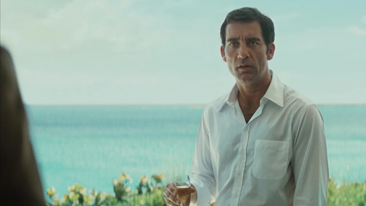 Duplicity (2009) movie still: medium — An outdoor terrace overlooks a bright turquoise sea under a clear daytime sky. A man in a white dress shirt…