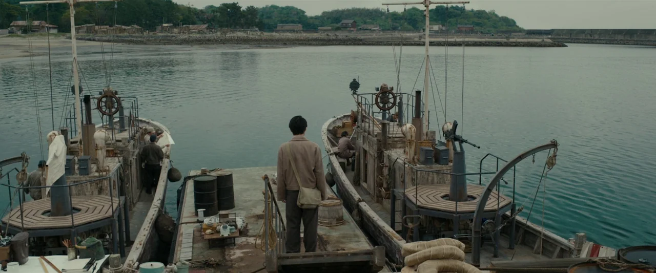 Godzilla Minus One (2023) movie still: wide — A man stands on a central concrete pier between two aged wooden transport vessels. The background…