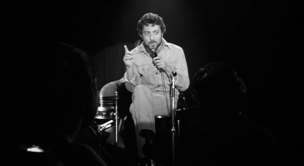 Lenny (1974) movie still: medium — A dimly lit nightclub features a performer illuminated by a single spotlight on set. The subject sits on a stool…
