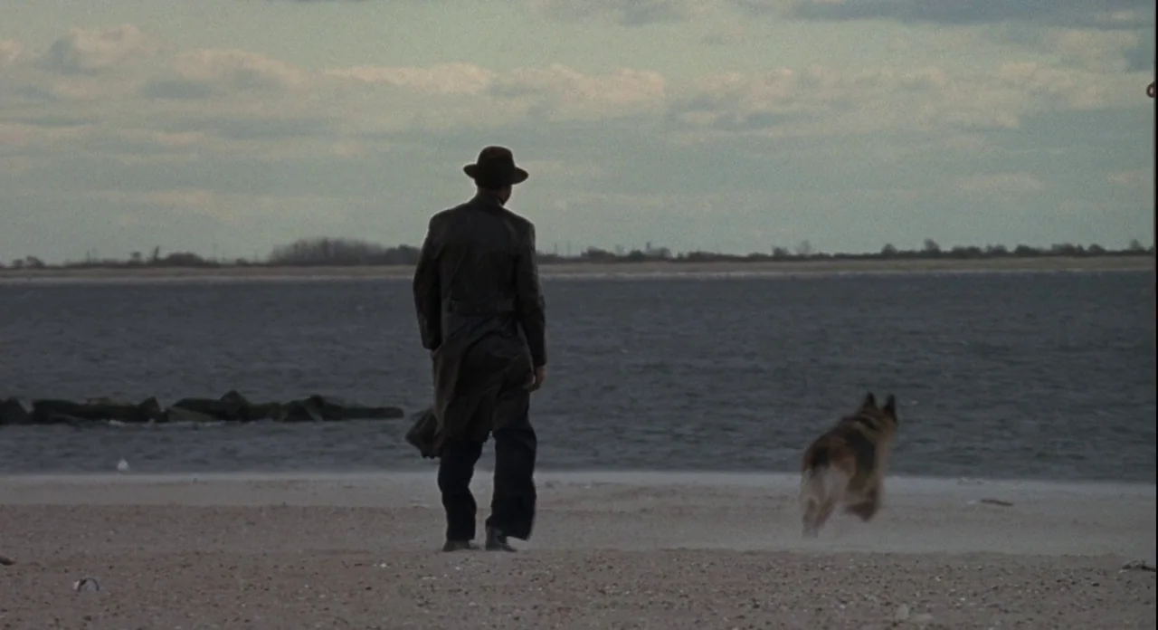 American Gangster (2007) movie still: fullbody — A man walks along a sandy shoreline by a body of water. An adult dog trots ahead of him near the…