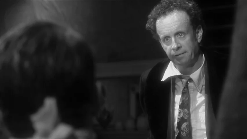 Keyhole (2011) movie still: medium — A dark indoor setting is framed from behind the shoulder of another person. The man in focus wears formal…