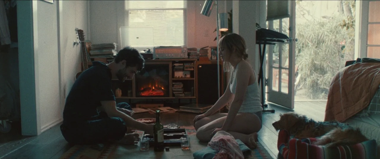 The Greatest Hits (2024) movie still: wide — A cluttered apartment room features an electric fireplace and scattered belongings on the floor. A man…