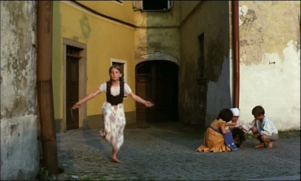 Woyzeck (1979) movie still: wide — A quiet cobblestone area sits between weathered buildings with peeling plaster and arched doorways. A young girl…