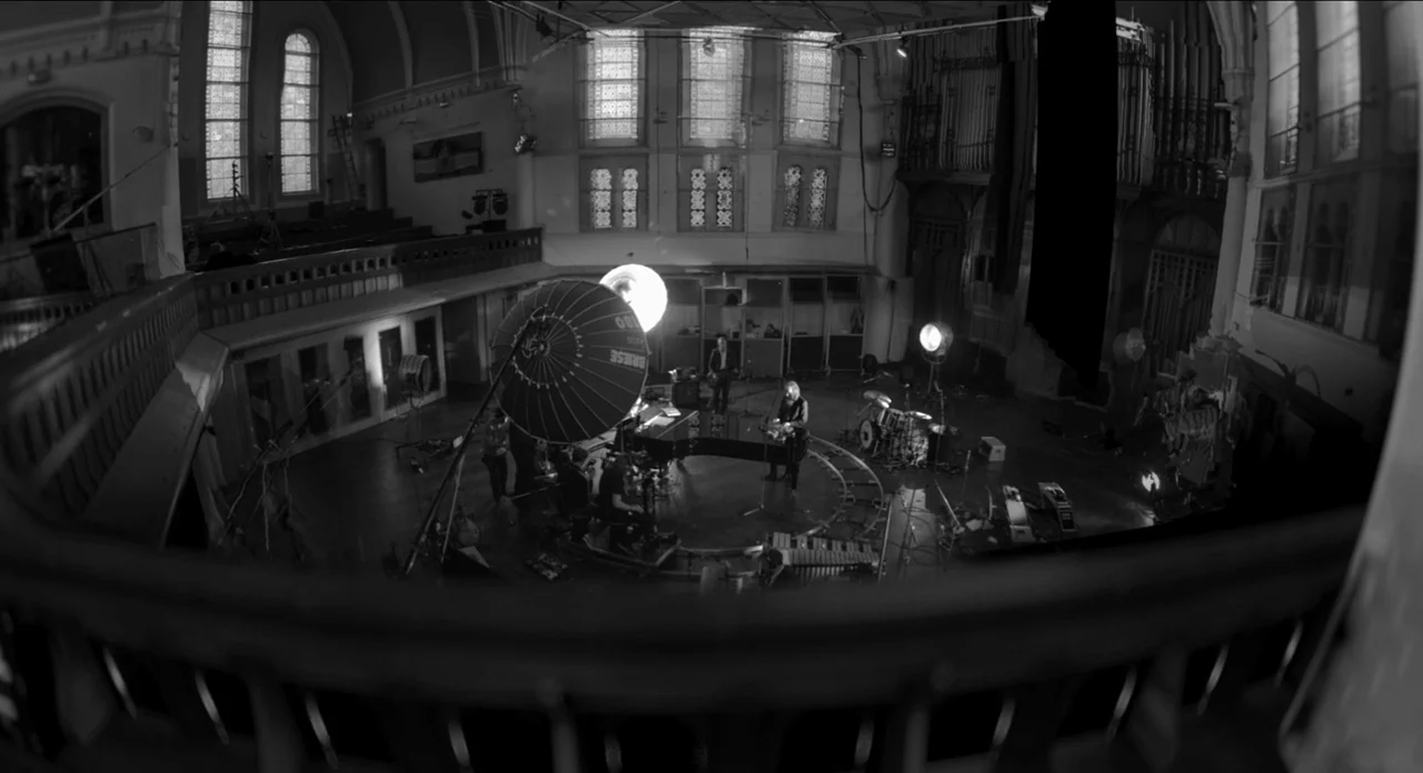 One More Time With Feeling movie still: wide — A large converted church hall with arched windows and a wooden interior floor. Musicians and crew…