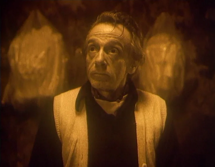 Dead Man’s Letters (1986) movie still: medium — A dimly lit, dark space features an elderly man standing before two plastic-wrapped objects. The man…