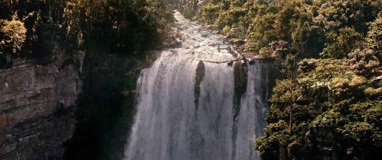 Tomb Raider (2018) movie still: establishing — A wide waterfall drops over a jagged rock ledge surrounded by dense forest foliage. Rushing water…