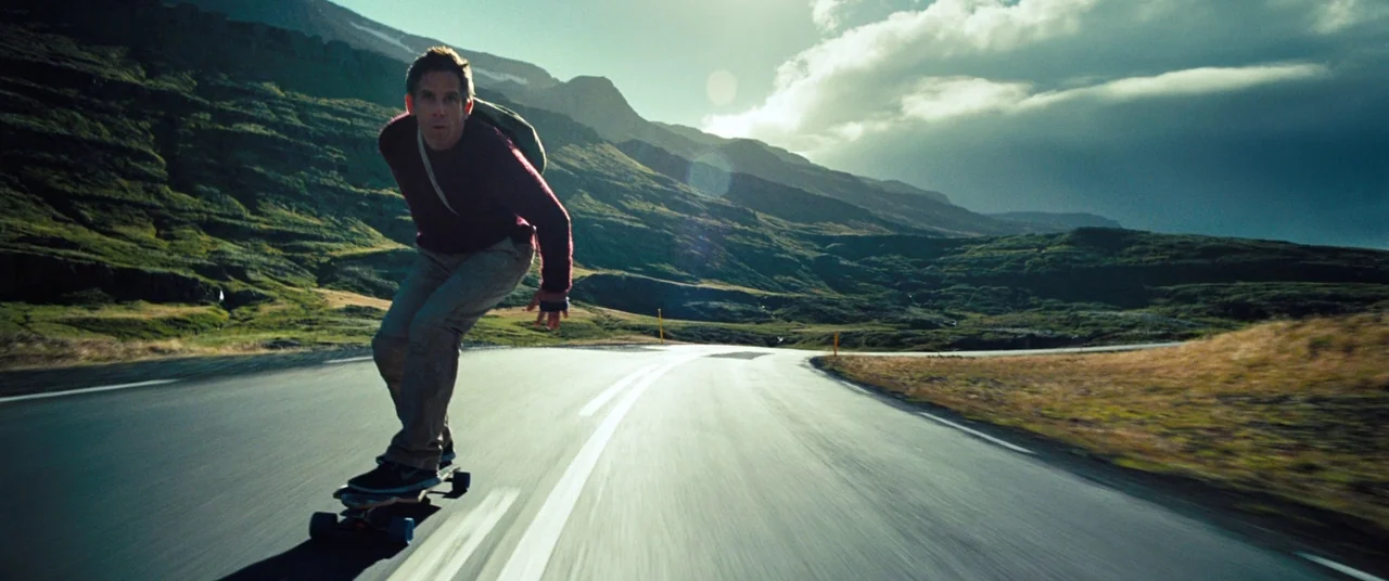 The Secret Life of Walter Mitty (2013) movie still: fullbody — A man skateboards along a smooth asphalt road cutting through vast, grassy mountain…