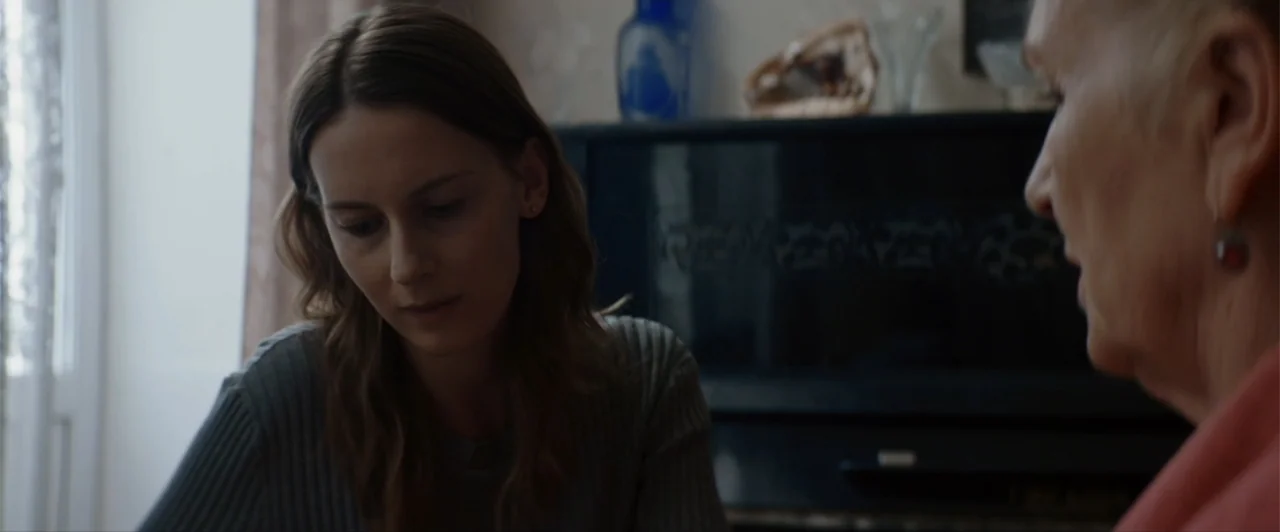 La herencia (2010) movie still: medium — A quiet indoor space features a dark piano placed against a patterned wall. A young woman listens…