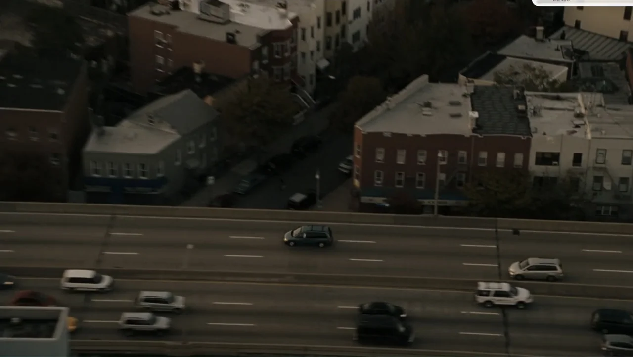 Fighting (2009) movie still: establishing — An aerial perspective shows a multi-lane highway running alongside dense urban brick buildings. Multiple…