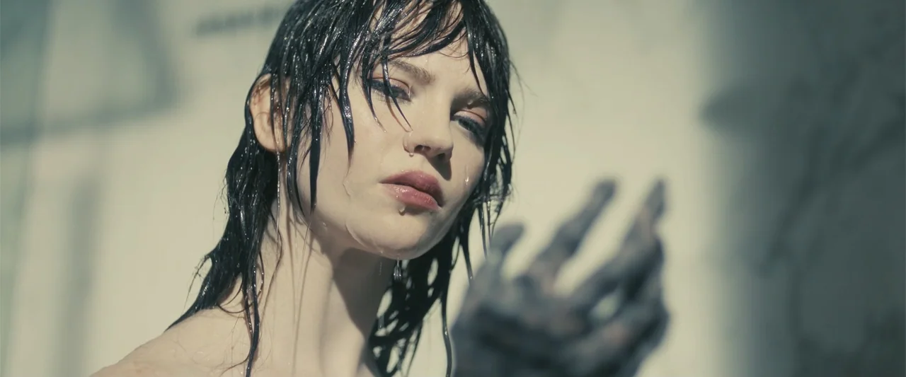 Companion (2025) movie still: closeup — A close-up against a light-colored wall with visible soft-focus shadow patterns. A woman with wet hair faces…