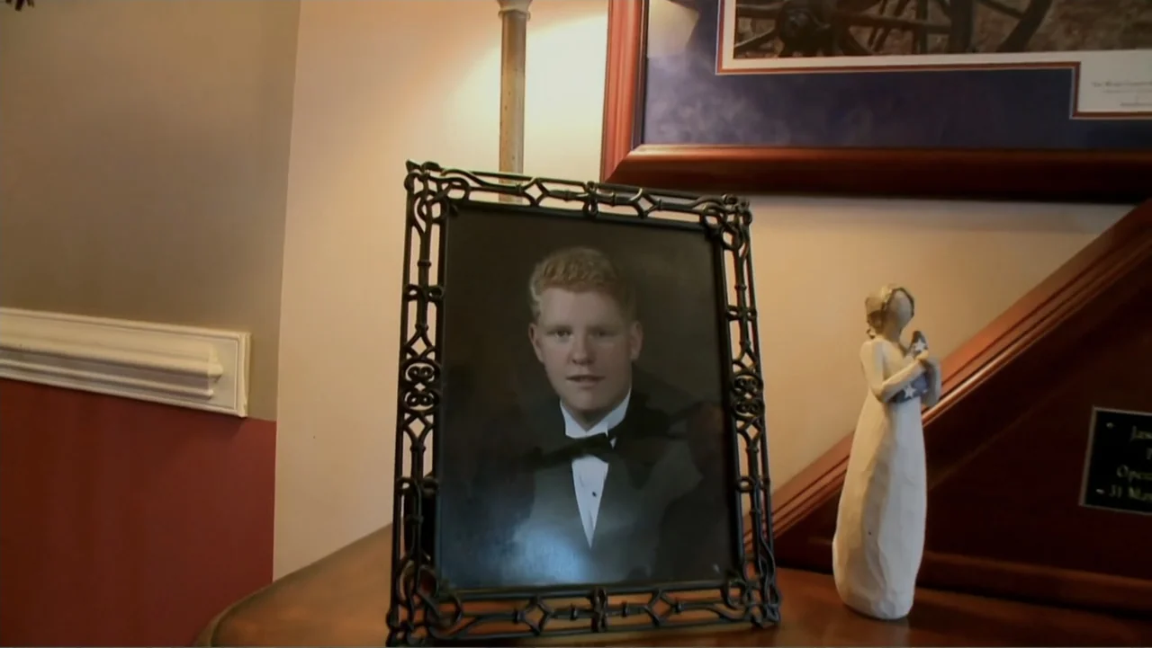 Wartorn: 1861-2010 (2010) movie still: closeup — A wooden shelf holds a framed portrait of a man in a tuxedo. A stylized abstract figurine stands…