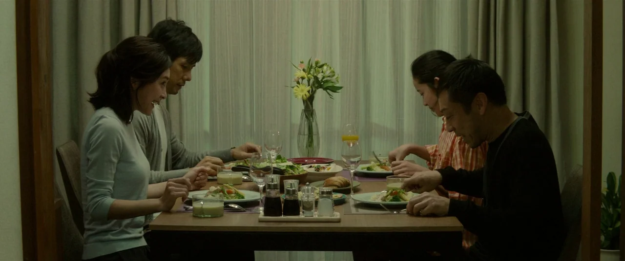 Creepy (2016) movie still: medium — A group of four adults and a teen sit around a wooden dining table. They eat dinner in front of a window covered…