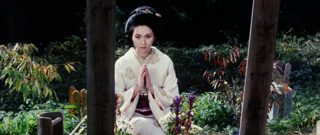 Lady Snowblood (1973) movie still: medium — A woman kneels among wooden headstone markers in a lush, green outdoor cemetery. She holds her hands…