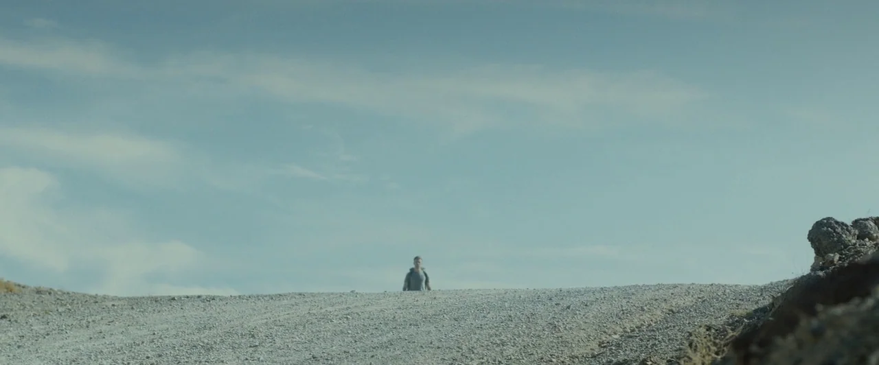 The Guest (2014) movie still: wide — A wide, barren gravel path crests against a pale blue sky filled with clouds. A lone man with a backpack walks…