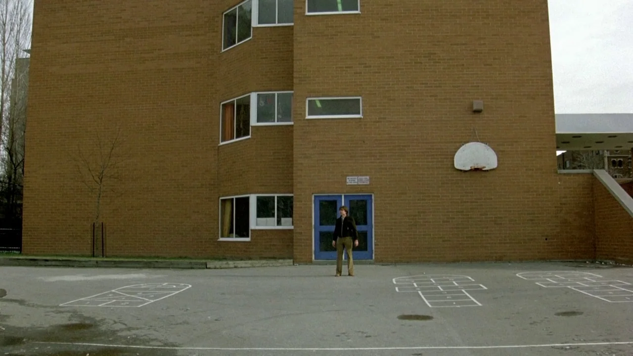 The Brood (1979) movie still: wide — A tan brick school building exterior features a blue double door and a basketball hoop. A lone young man stands…
