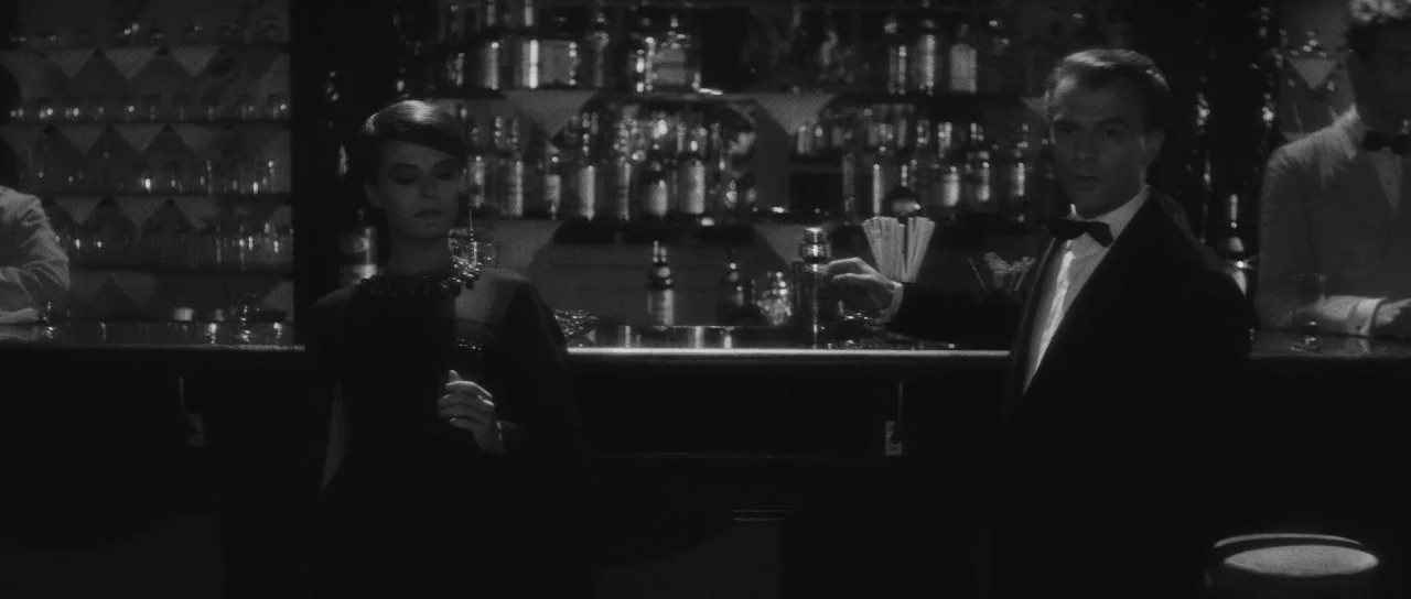Last Year at Marienbad (1961) movie still: medium — A dimly lit lounge featuring a bar stocked with many glass bottles. A man and a woman in formal…