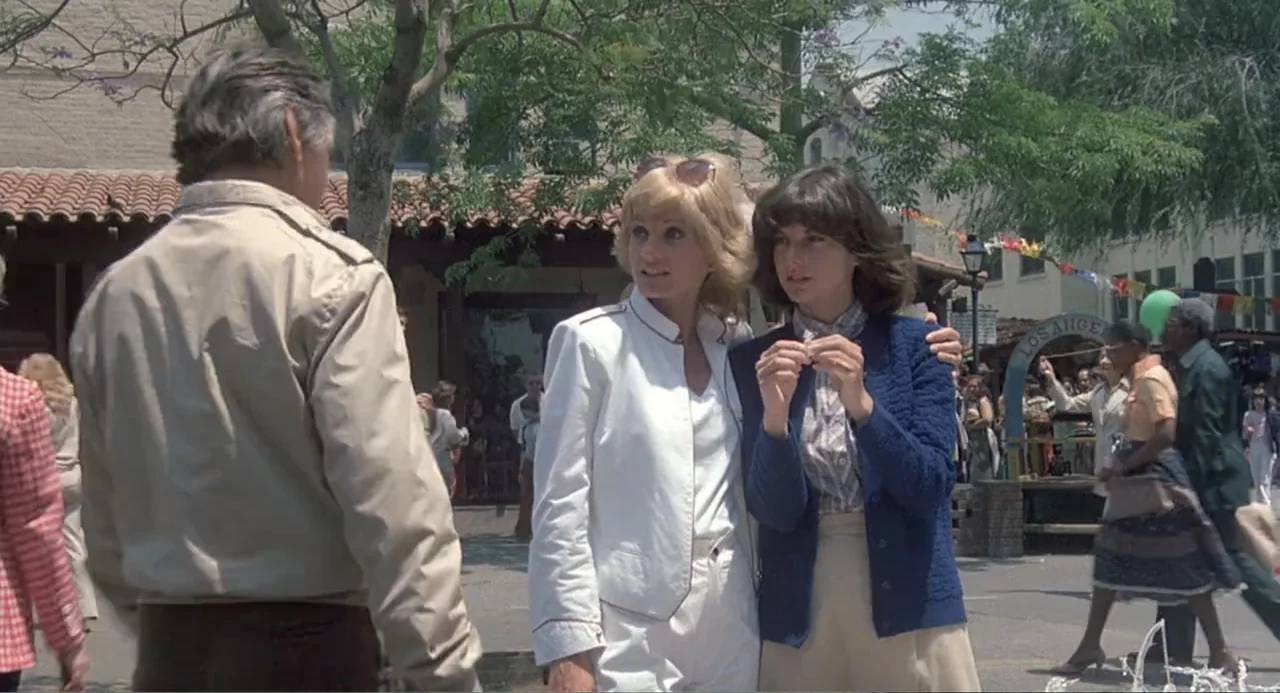 Death Wish II (1982) movie still: medium — A sunny public plaza features terracotta tiled roofs and trees under a clear sky. Three people stand in…