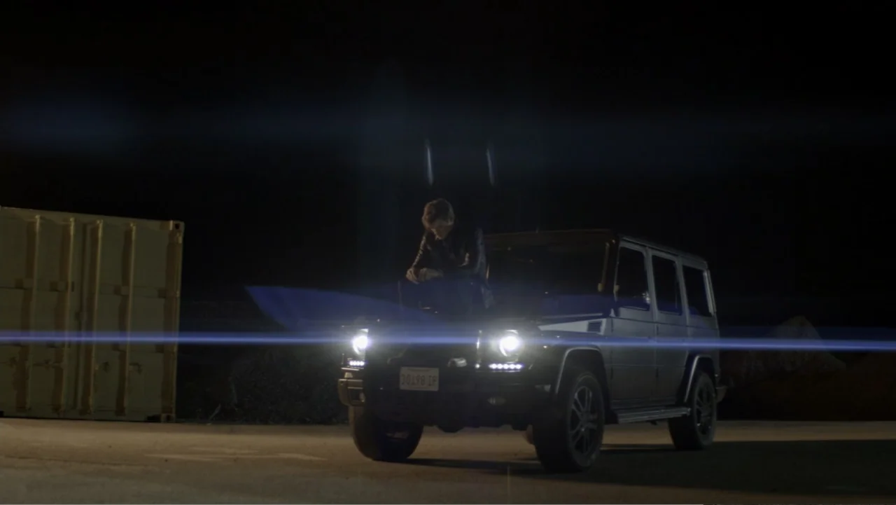The Collective (2023) movie still: wide — An empty industrial lot at night contains a large shipping container and dark pavement. A man sits…