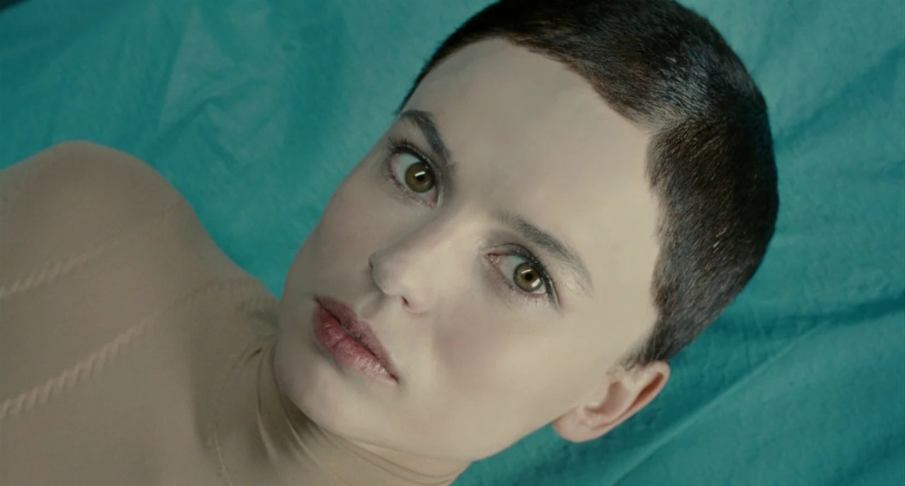 The Skin I Live In (2011) movie still: closeup — A woman with short dark hair lies on a teal-colored medical surface. She looks directly upward with…