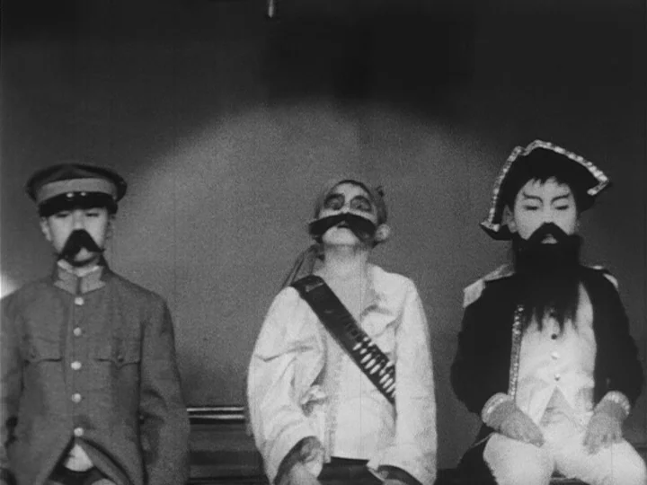 Emperor Tomato Ketchup movie still: medium — A bare stage with a flat gray wall serves as the background. Three young children wearing different…