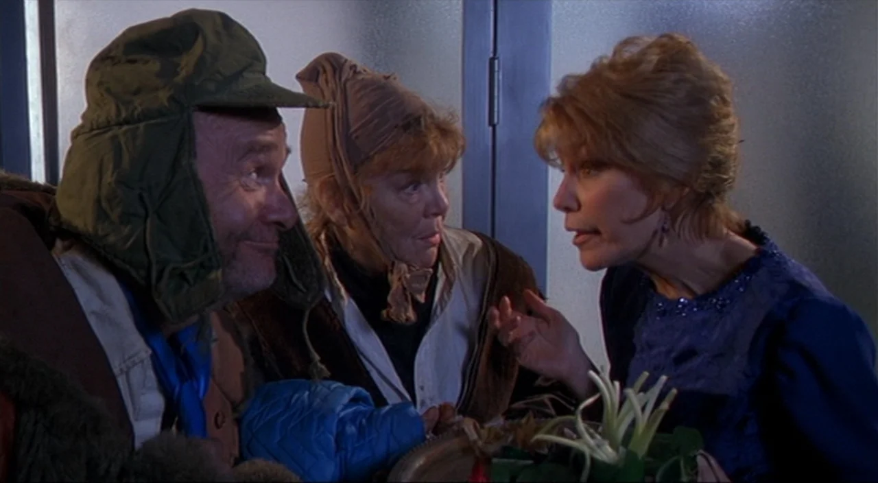 Pecker movie still: medium — A dimly lit room with three people engaged in a discussion. An older man in a hunter hat stands near a woman wearing a…