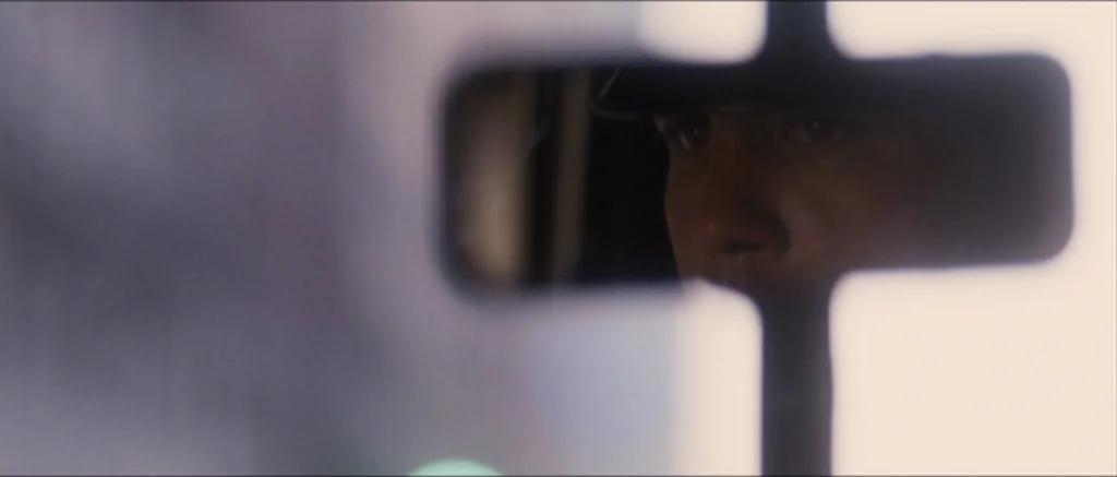 The Killer Inside Me (2010) movie still: closeup — A man is viewed through a small, rectangular gap in a white partition. The subject is dimly lit…