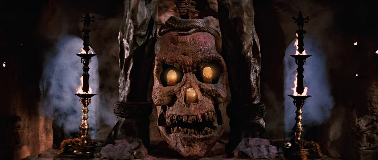 Indiana Jones and the Temple of Doom (1984) movie still: wide — A dark stone interior features a large, weathered skull sculpture resting on a…