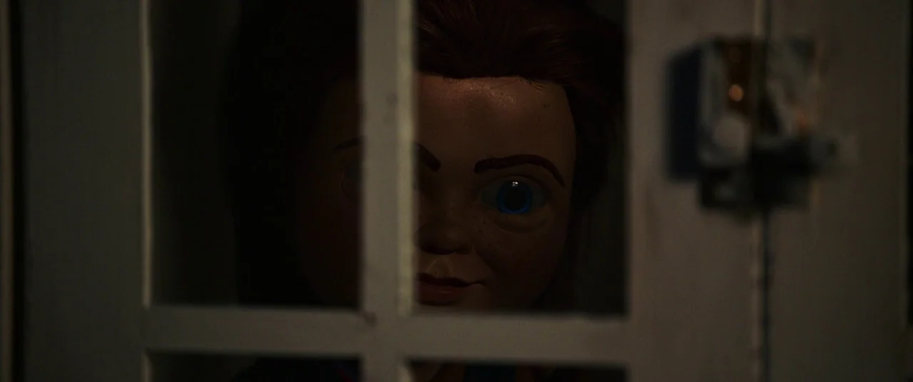 Child's Play (2019) movie still: closeup — A doll face is framed behind the white wooden bars of a glass door. The figure watches from the interior…