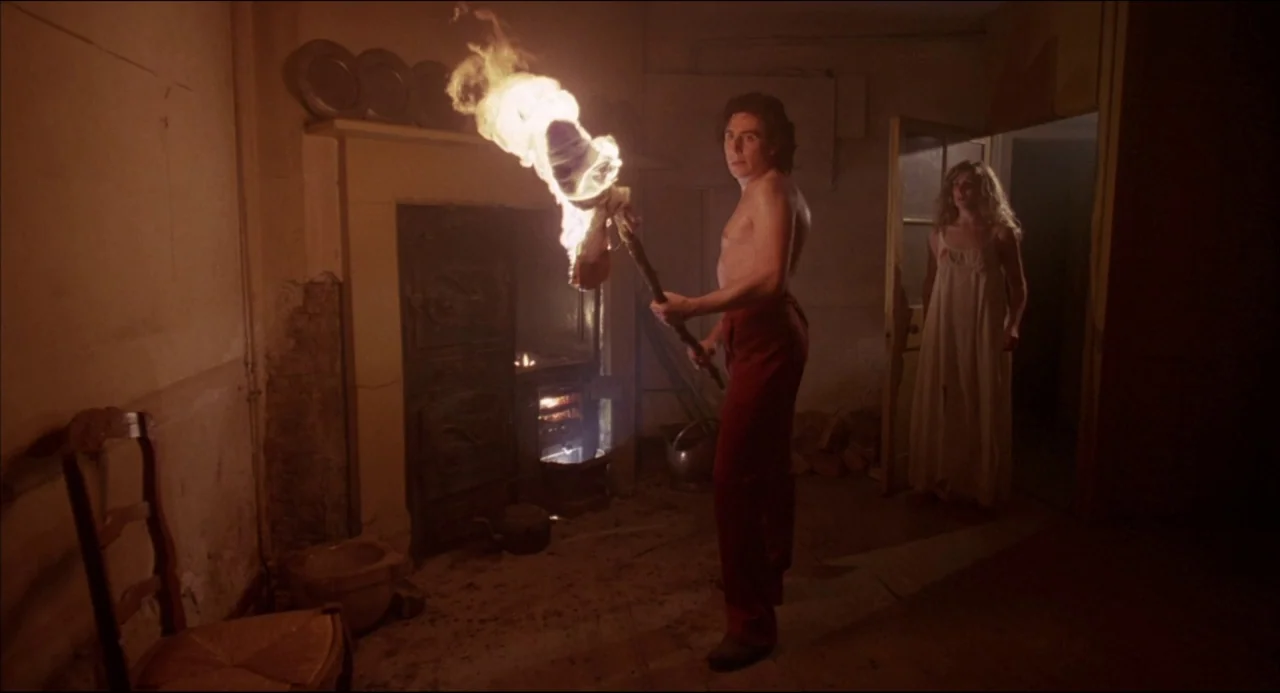 Gothic (1986) movie still: medium — A dark, dilapidated room contains a fireplace with glowing embers and a wooden chair. A shirtless man holds a…
