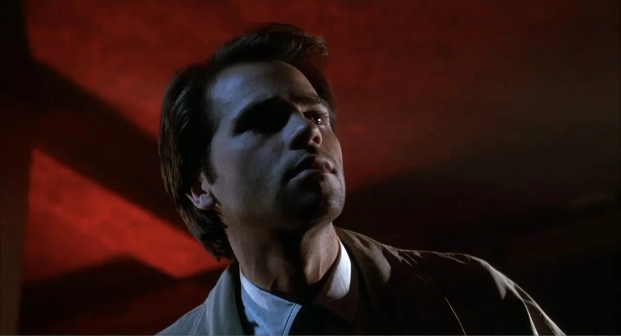 I, Madman (1989) movie still: closeup — A dimly lit room features dark red walls illuminated by a harsh light source. A man in a tan trench coat…