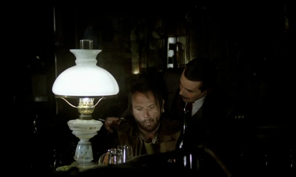 Despair (1978) movie still: medium — A dimly lit bar interior features a glowing glass lamp on a wooden counter. A man in a dark suit leans toward a…