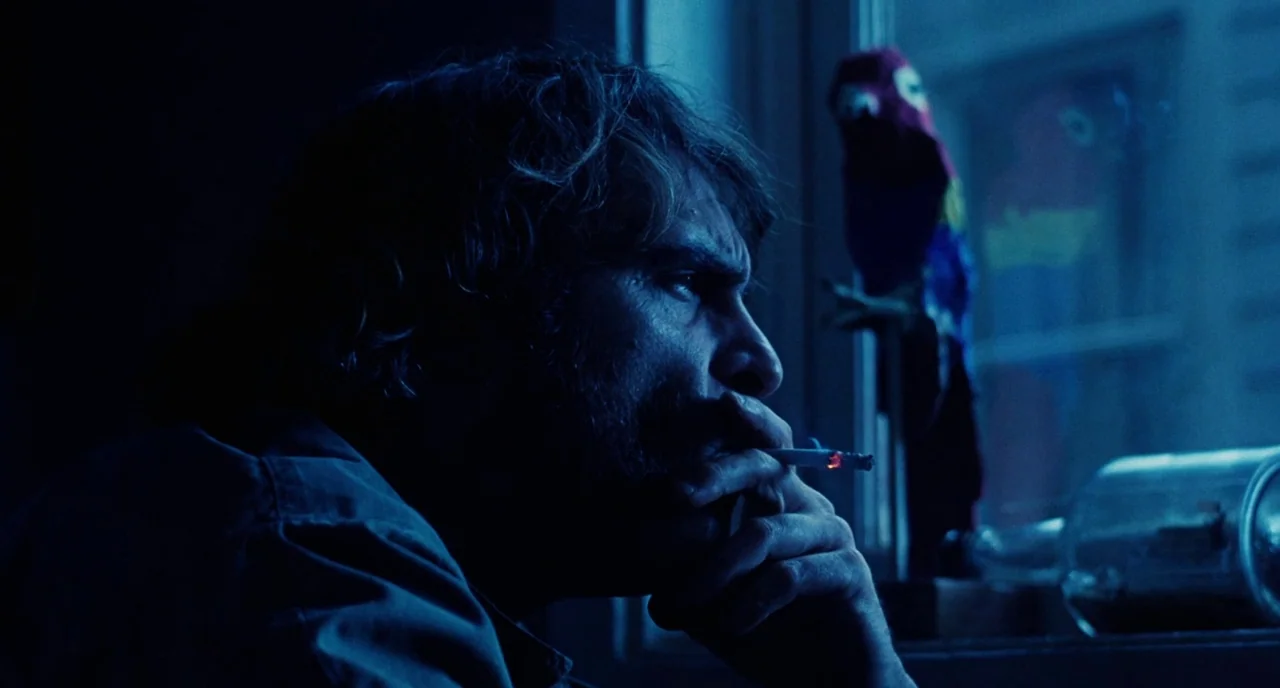 Inherent Vice (2014) movie still: closeup — A dimly lit room at night with a blue-hued window frame visible in the background. A bearded man sits by…