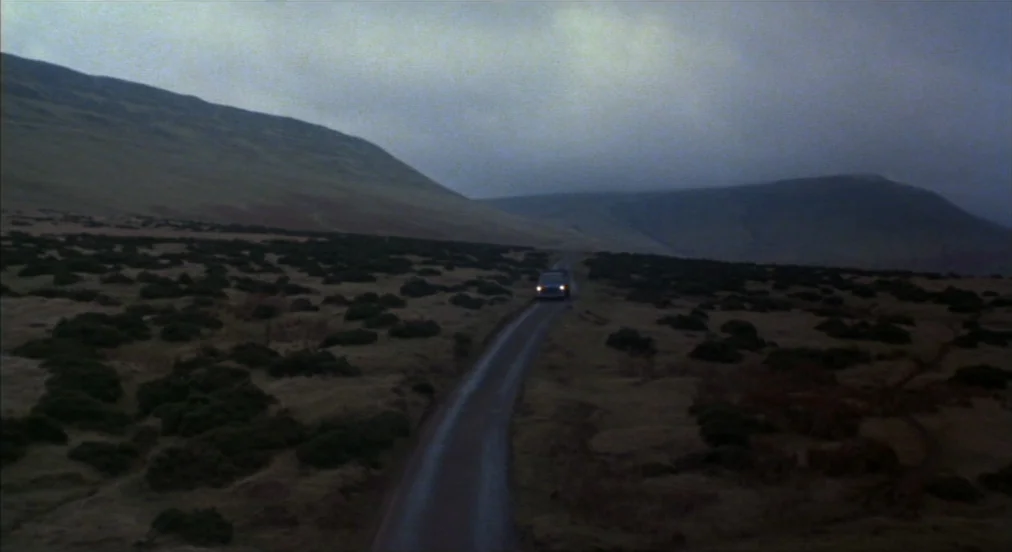 An American Werewolf in London (1981) movie still: establishing — A remote single-lane asphalt road winds through a wide, heather-covered moorland…