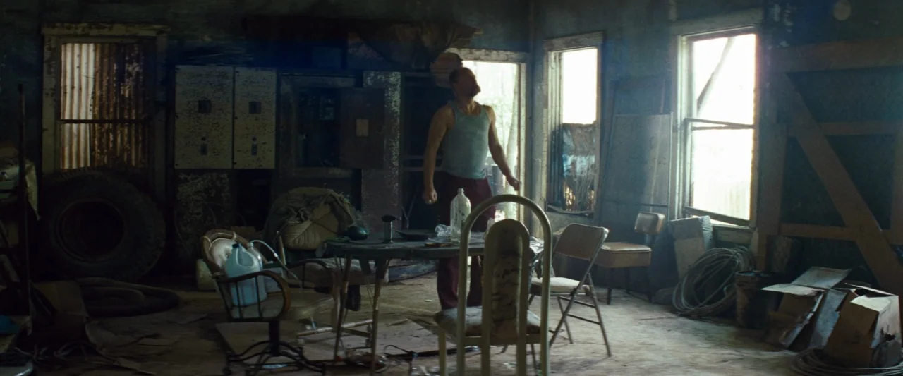 Out of the Furnace (2013) movie still: fullbody — A dilapidated room filled with assorted junk, chairs, a table, and tires. A lone man stands in the…