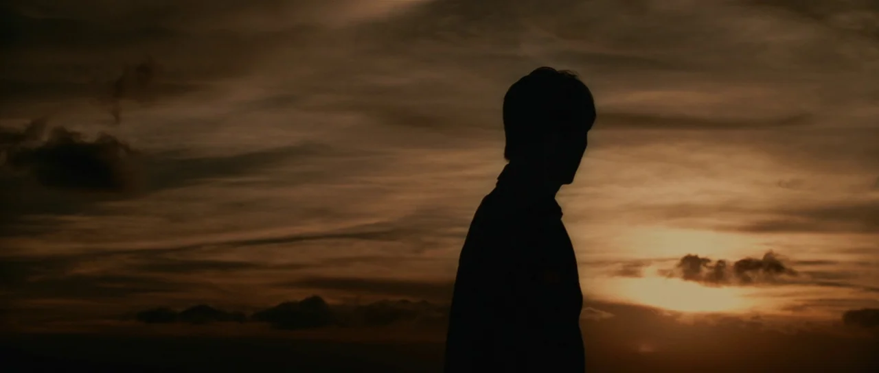 Infernal Affairs (2002) movie still: medium — A dark figure stands against a sprawling, amber-lit sky filled with textured clouds. The person…
