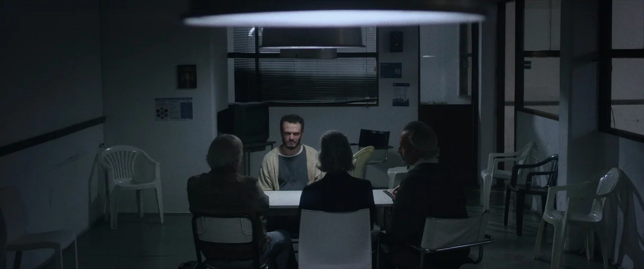 Terrified (2017) movie still: wide — A sparse meeting room contains four people seated around a central table. The walls are bare and punctuated by…